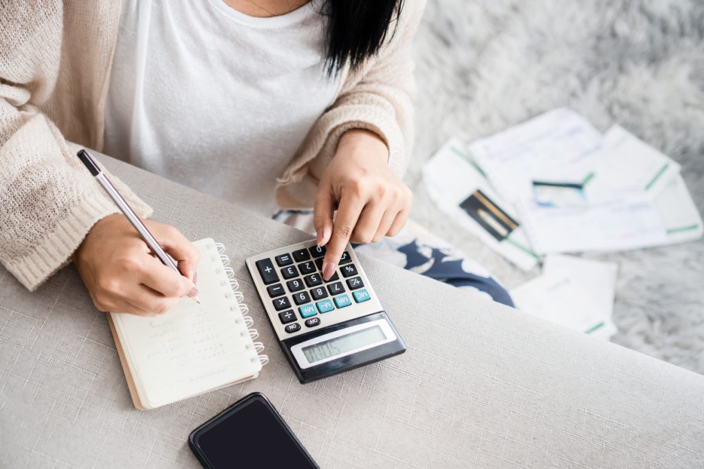 Woman writing a list of finances on notebook calculating her expenses with calculator and many invoices
