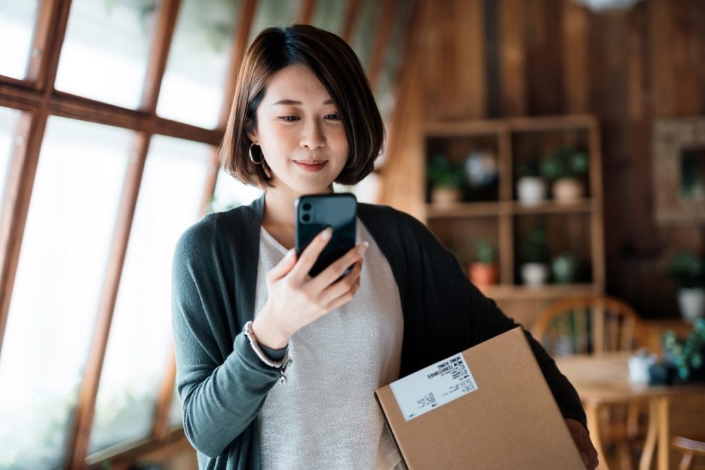 Smiling woman shopping on eCommerce website with smartphone on hand, receiving a delivered parcel by home delivery service