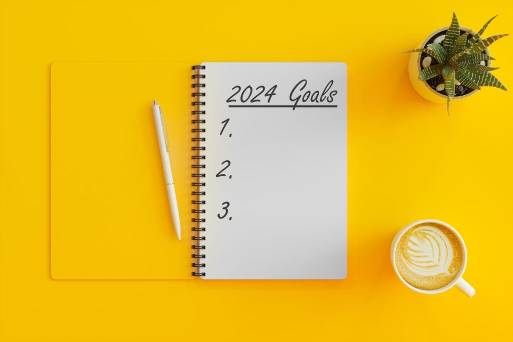 Notebook with 2024 goals written on it on a yellow table with a coffee and plant. Notebook to be used for digital marketing goals exercise. 