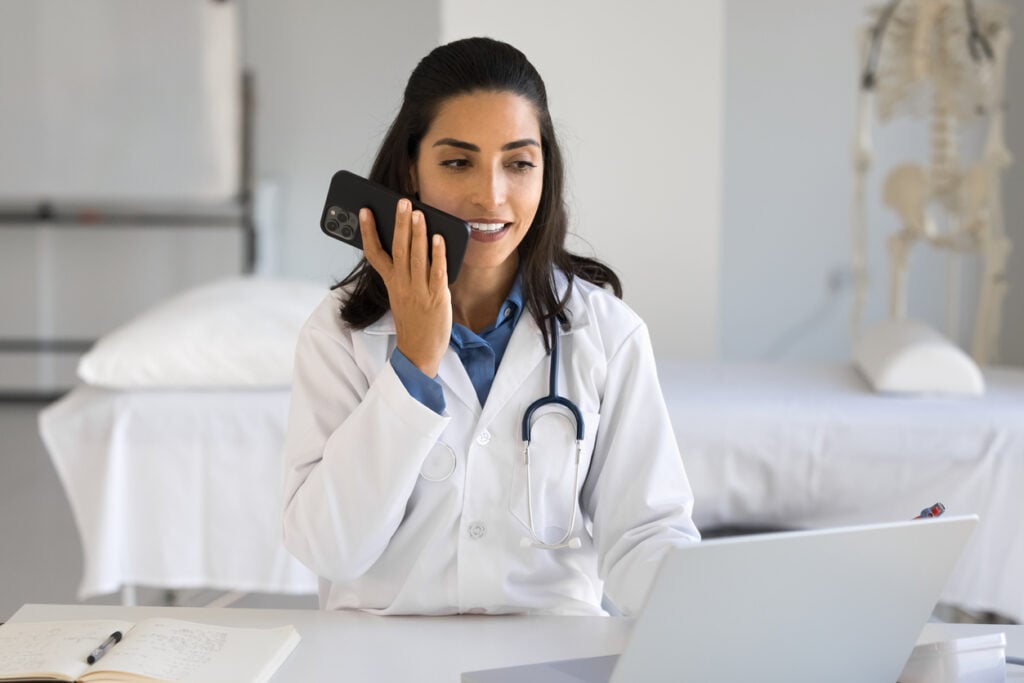 Female doctor in a lab coat using voice search on her phone while working at a desk in a medical office.