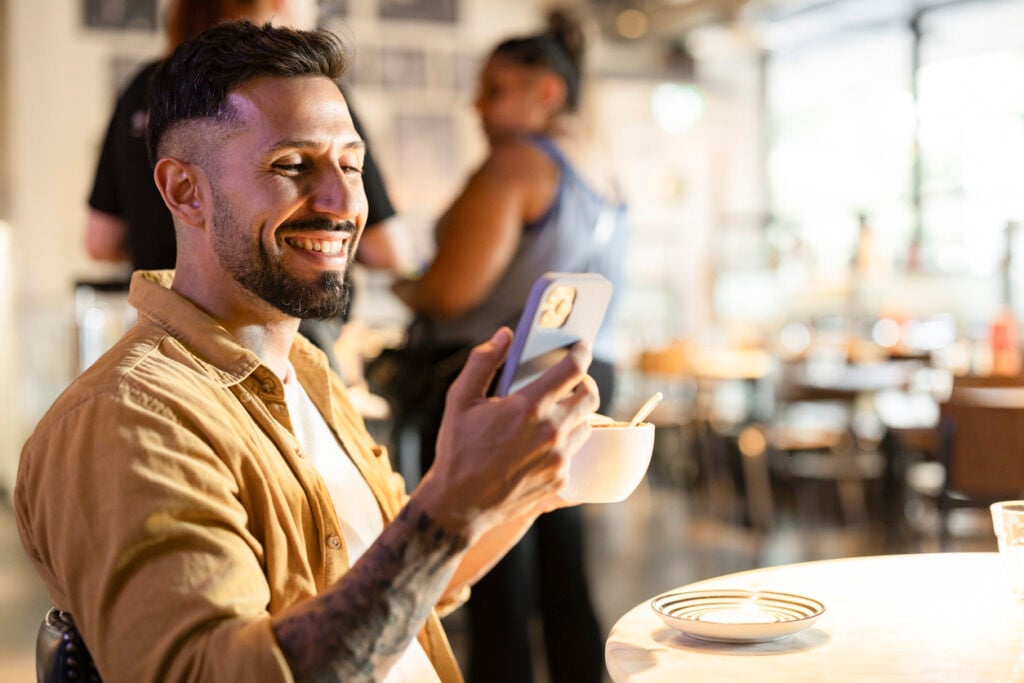 Smiling man using voice search on his smartphone while enjoying coffee in a lively café.